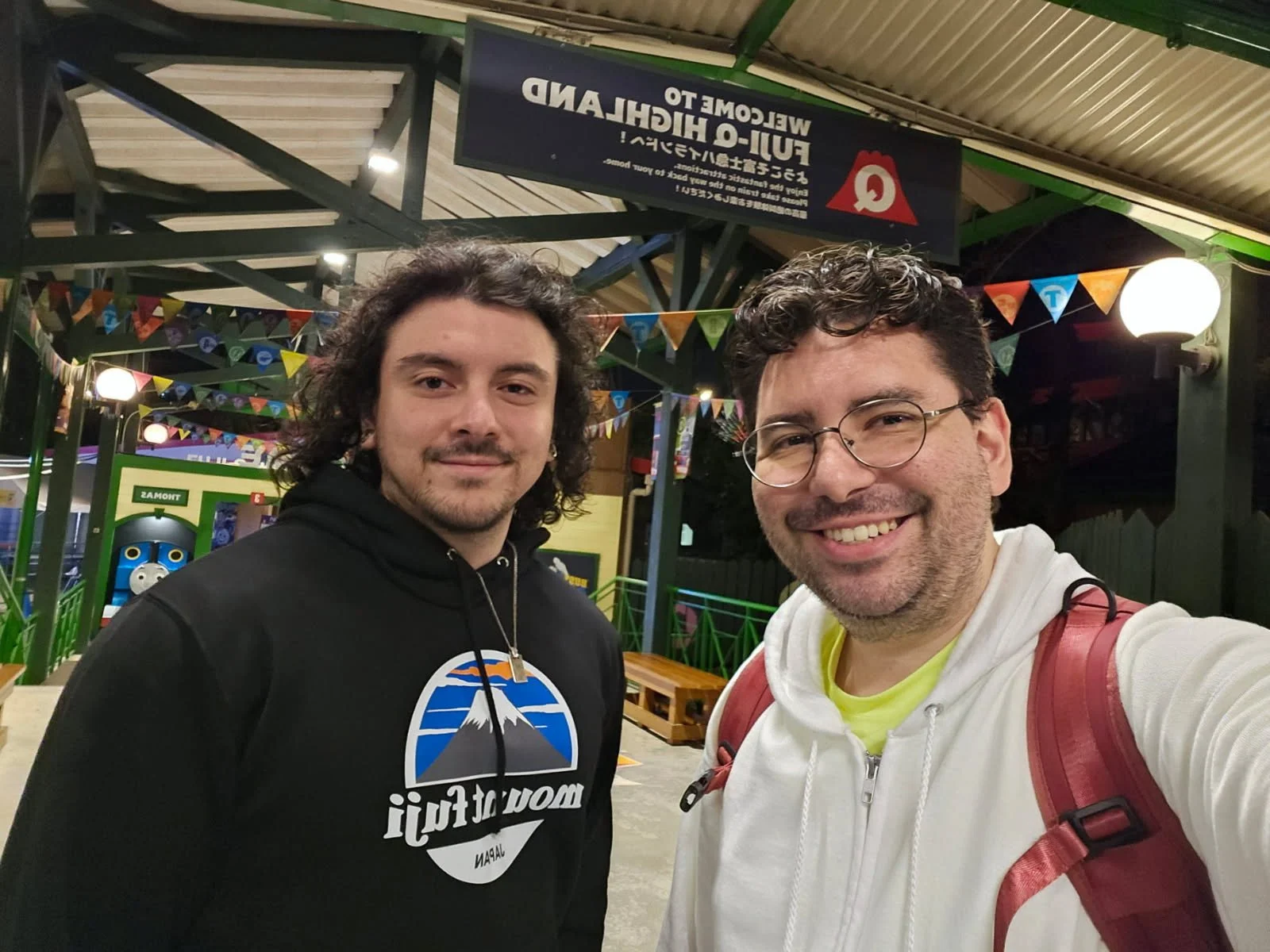 Two men smiling for a selfie at an amusement park or carnival, with colorful flags and a train-themed attraction in the background.