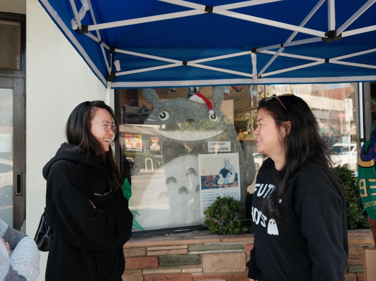 Two women smiling and talking in front of a glass window with a Totoro figure wearing a Santa hat behind it, under a blue canopy.