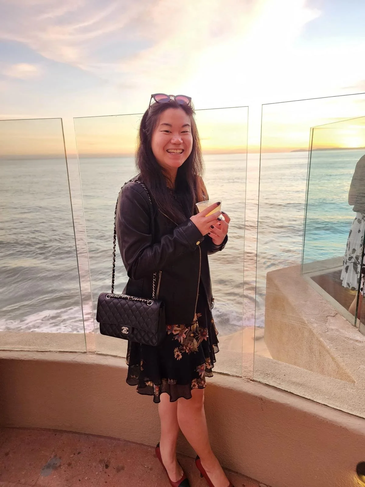 A woman smiling and holding a drink at sunset by the ocean, wearing a black jacket, floral skirt, and red shoes, with sunglasses on her head and a black quilted handbag.