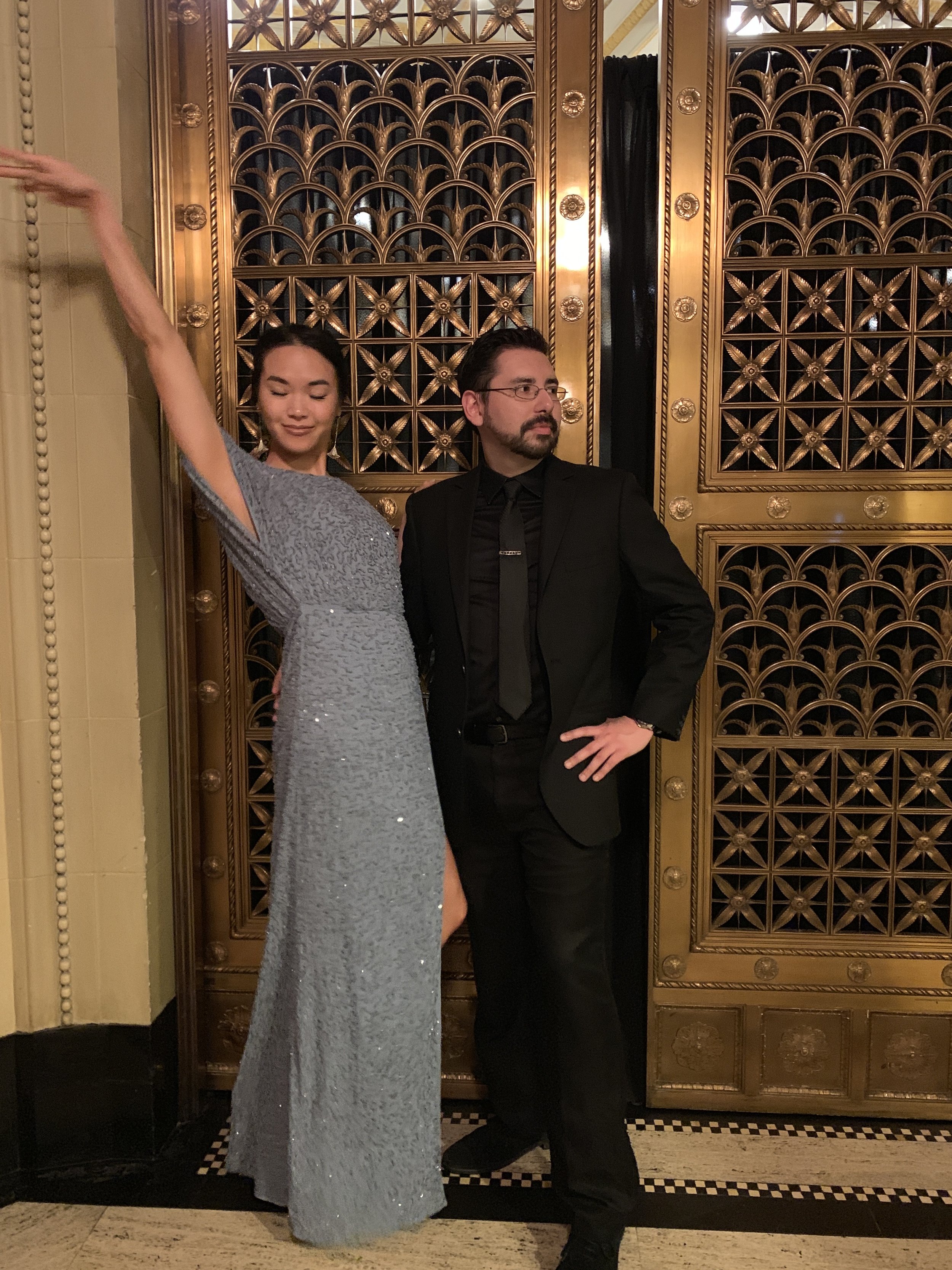 A woman in a long, silver, sequined gown stands with her eyes closed, arm raised, next to a man in a black suit with a black shirt and tie, in front of an ornate gold decorative door with intricate patterns.