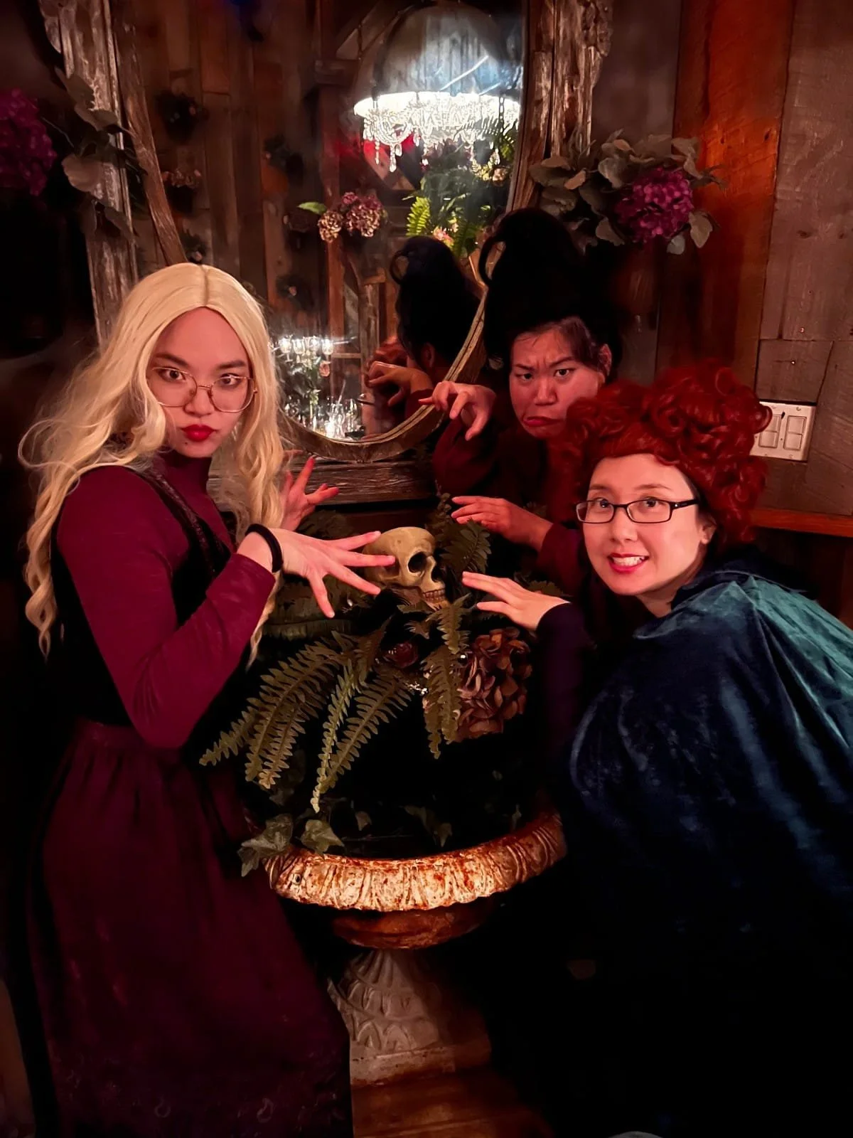 Four women dressed in Halloween costumes posing around a table with a small human skull, foliage, and a mirror reflecting their faces in a rustic, wood-paneled room.