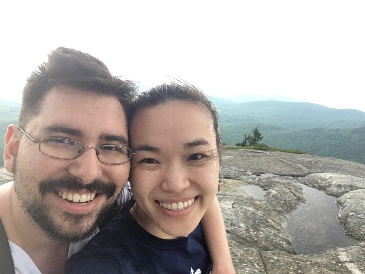 Smiling couple taking a selfie on a rocky mountain top with a mountain landscape in the background.