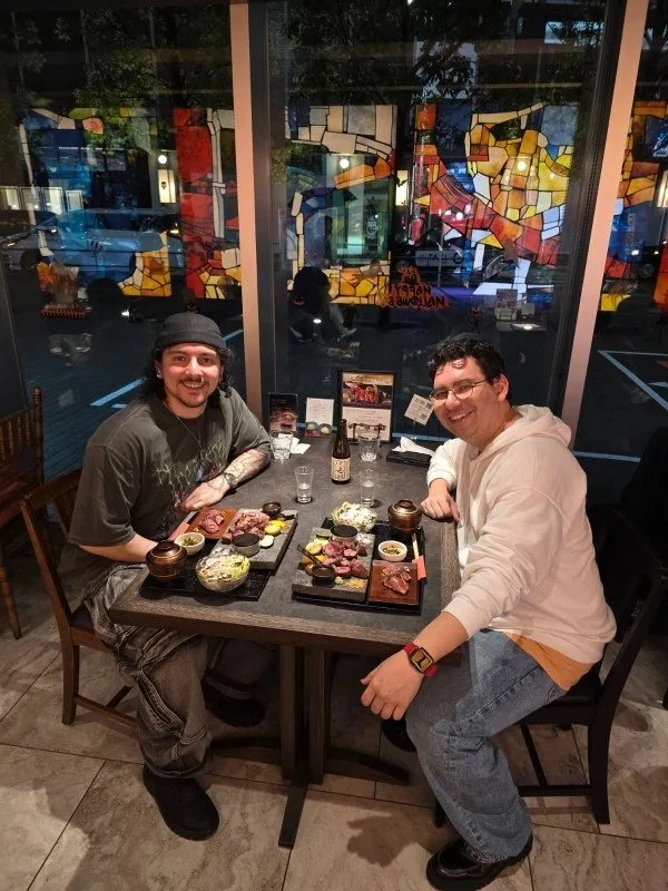 Two men enjoying a meal at a restaurant table with various dishes including sushi and grilled meats, sitting by a large window with colorful stained glass artwork in the background.