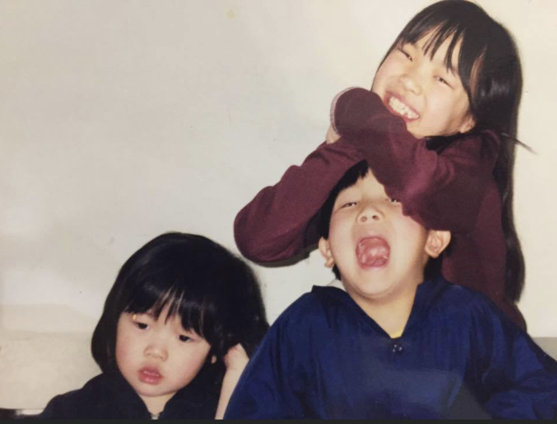 Three children with black hair, two girls and one boy, sitting together indoors. The girl at the back is smiling and hugging the girl at the front, who has a neutral expression. The boy is sitting in front, with his mouth open wide as if surprised or