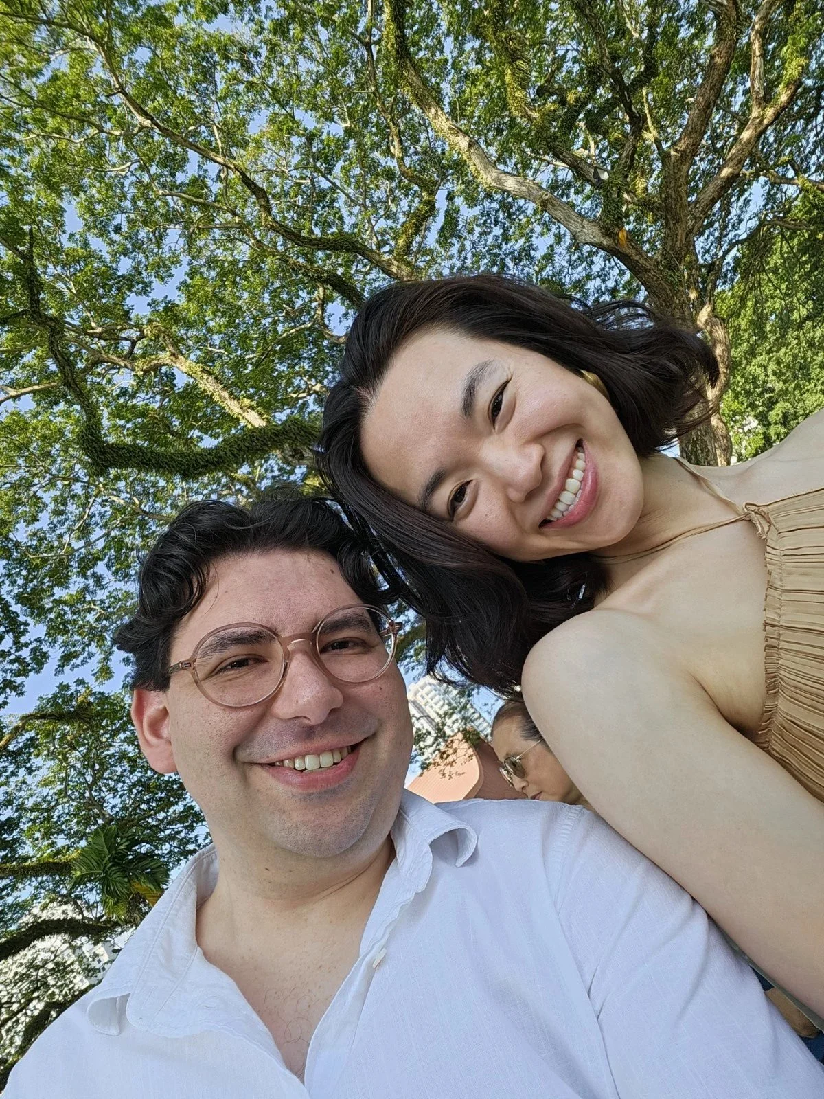 Two people taking a selfie outdoors under a large leafy tree, smiling at the camera, with bright blue sky visible through the branches.