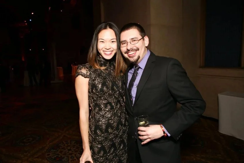A smiling woman and man posing together at an indoor event, the woman wearing a black lace dress and the man in a black suit with a purple shirt and tie, holding a drink.