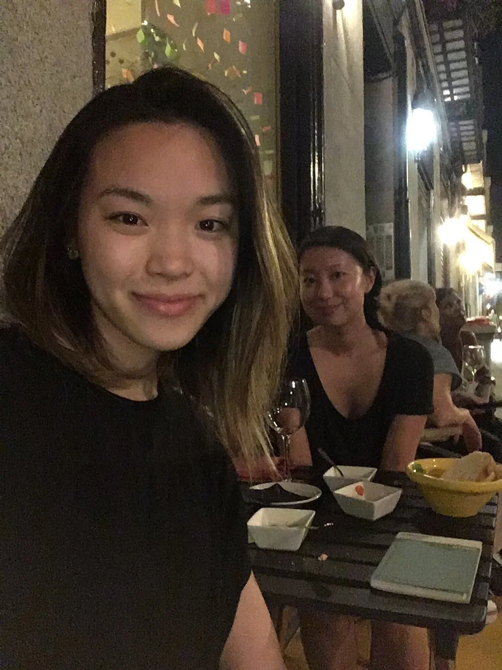 Two women seated at a table outside at night, with food and drinks, smiling at the camera. One woman in the foreground has shoulder-length hair, and the other woman behind her has dark hair and is wearing a black top.