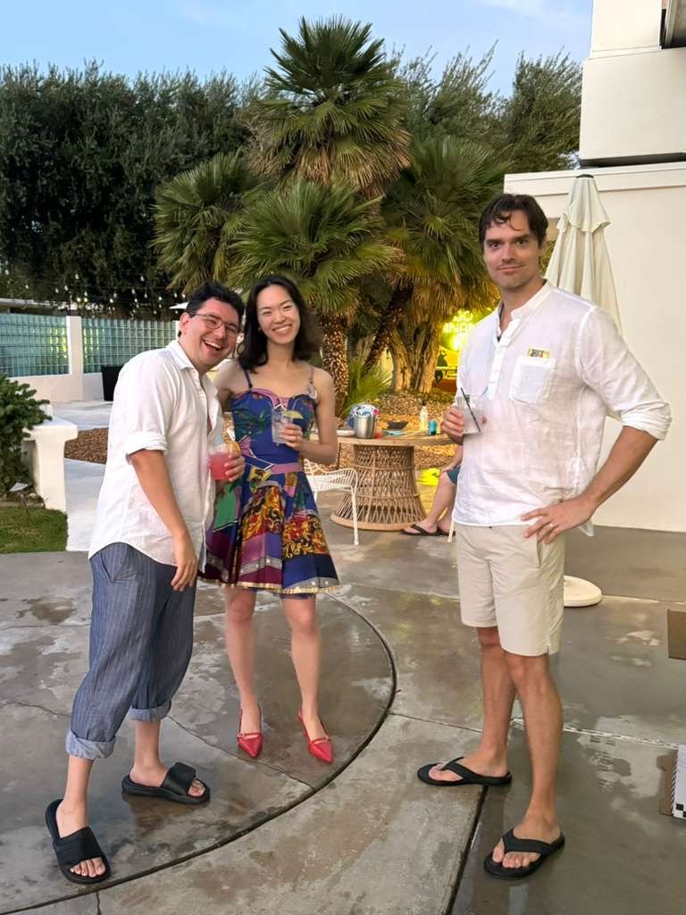 Three people enjoying drinks at a backyard party, standing on a wet concrete surface with palm trees and outdoor furniture in the background.