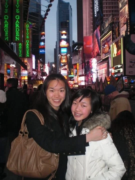 Two smiling young women hugging in Times Square at night, surrounded by bright neon billboards and a crowd of people.