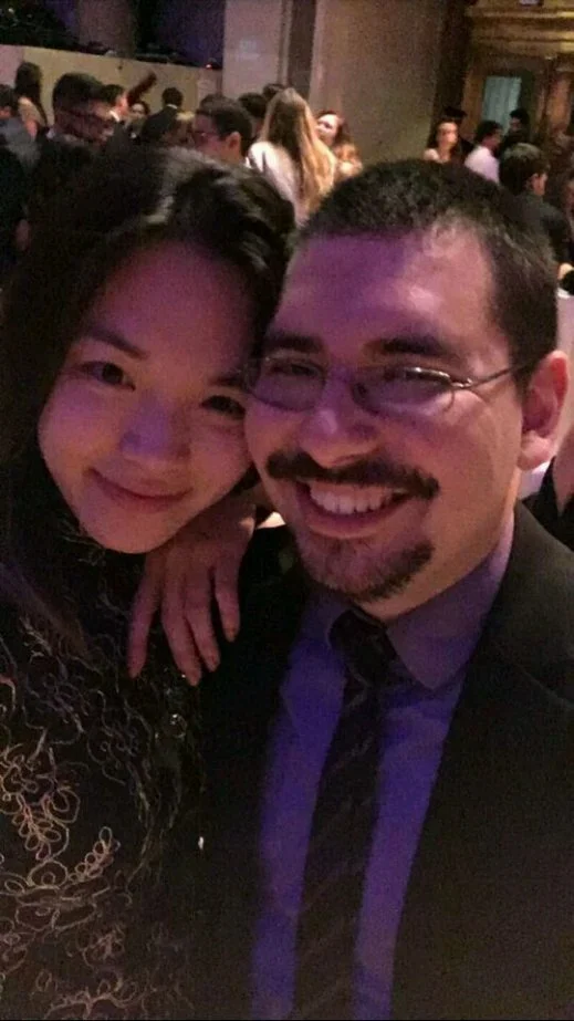 A selfie of a woman with long dark hair and a man with glasses and a mustache, both smiling, at a crowded event with many people in the background.