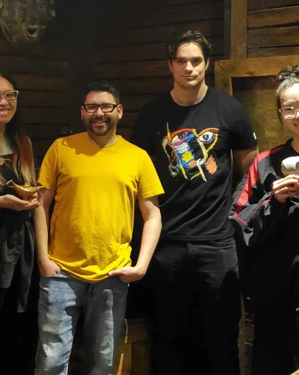 Group of four people standing together indoors, with wooden walls in the background. The second person from the left is wearing a yellow t-shirt and glasses, smiling. The third person is a tall man wearing a black t-shirt with a colorful face design.