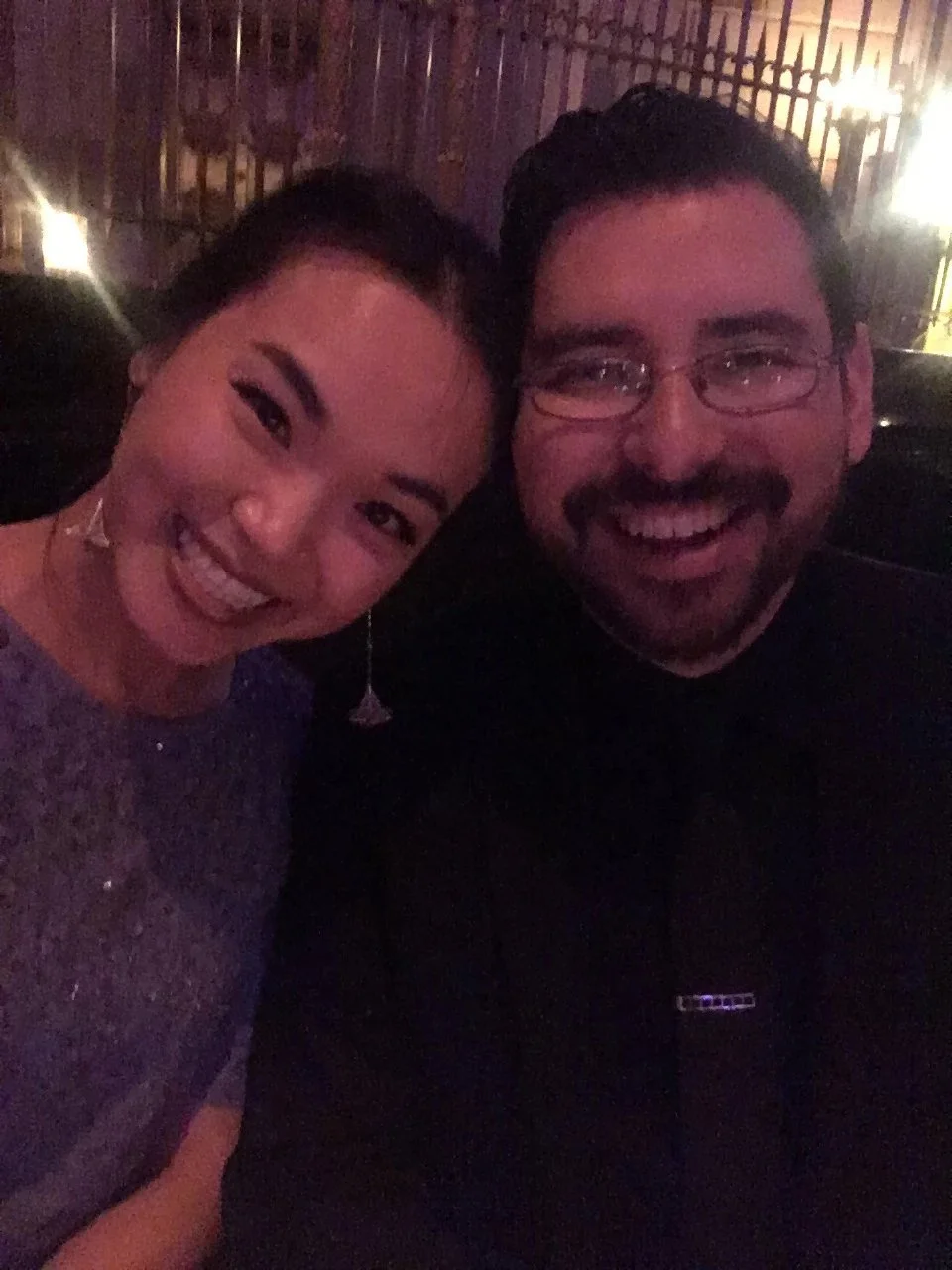 A woman and a man smiling and posing for a photo indoors at night, with dim lighting and a wooden lattice background.