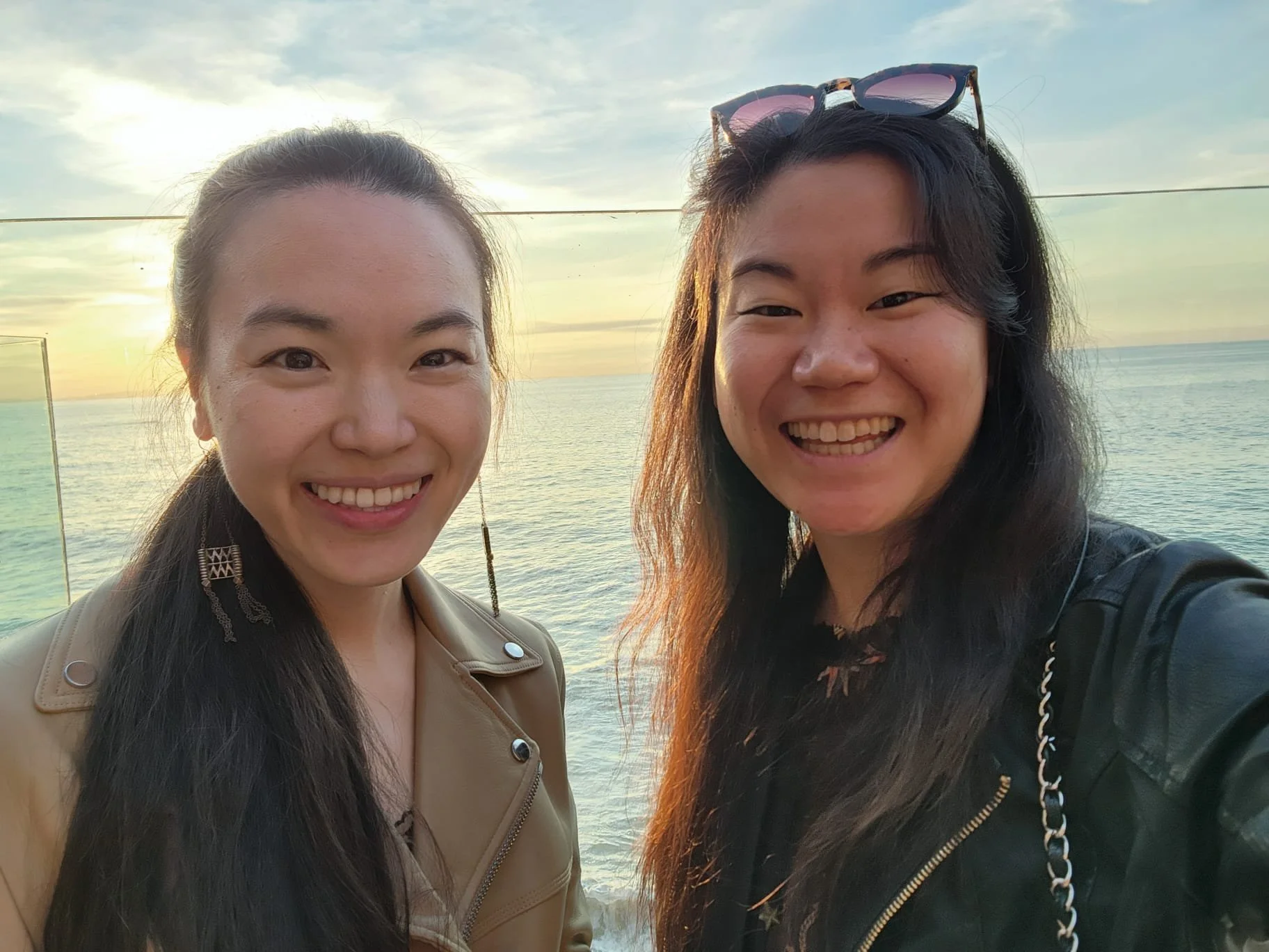 Two women smiling for a selfie at the beach during sunset, with the ocean and sky in the background.