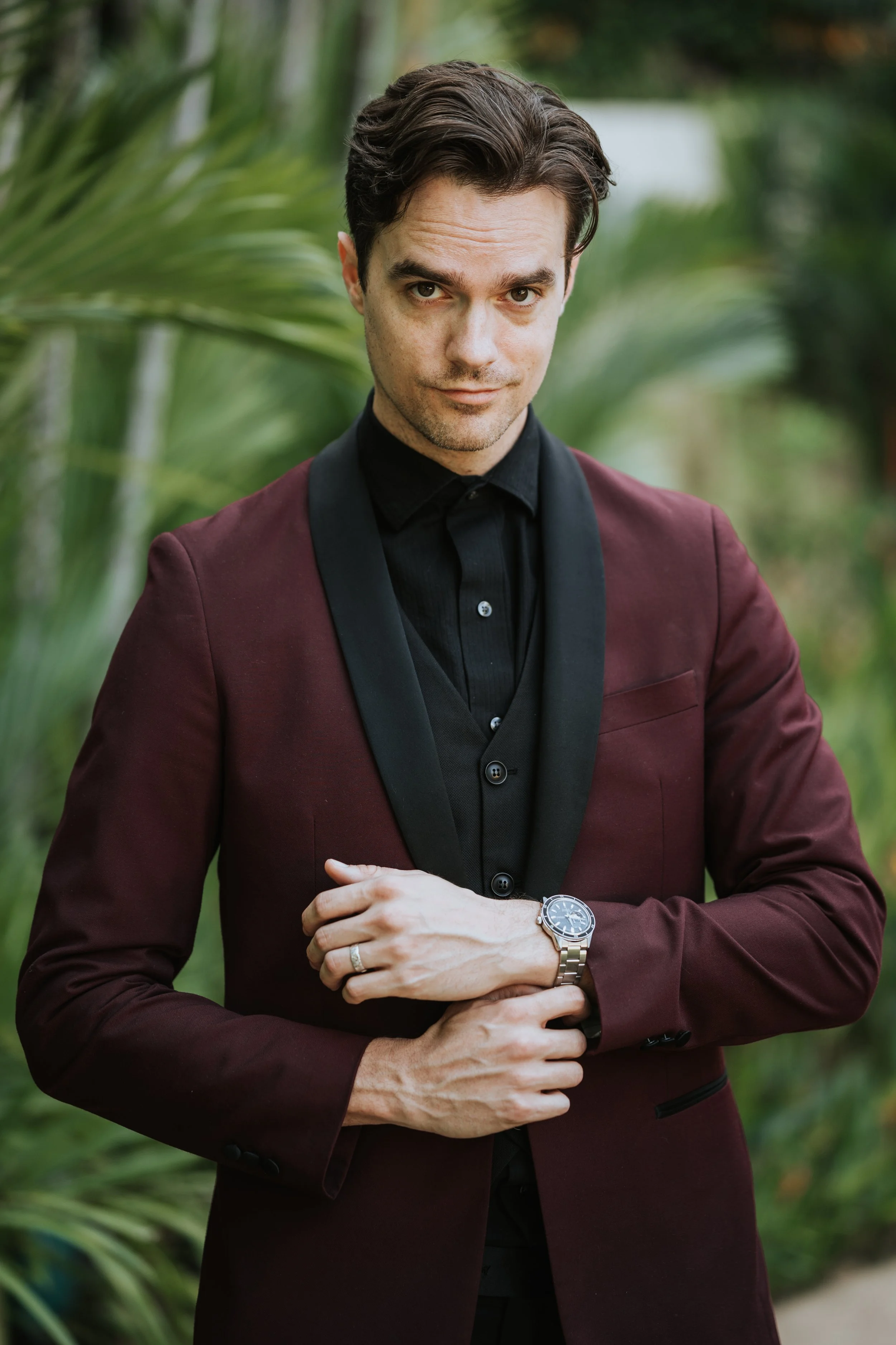 Man in burgundy blazer with black lapel and black shirt standing outdoors among greenery, wearing a silver watch and a wedding ring, looking at the camera.