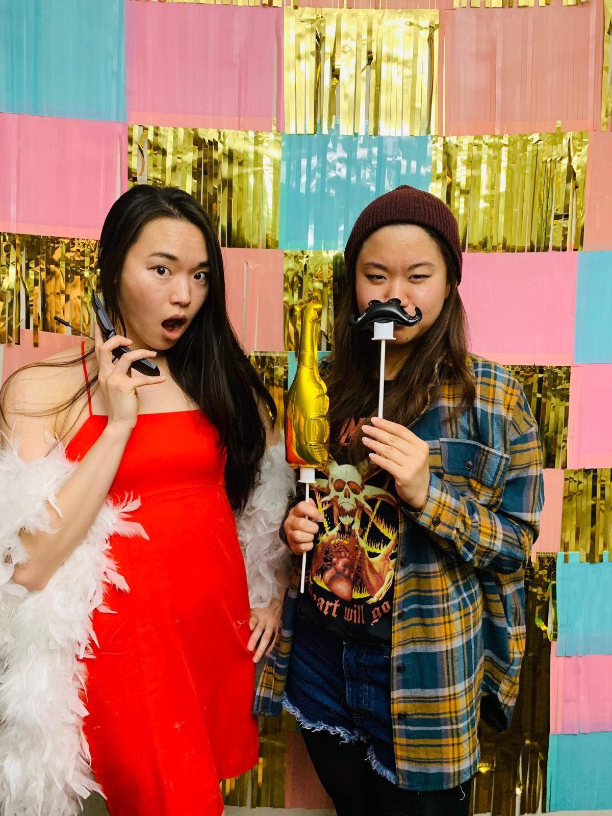 Two women celebrating at a party with pink, blue, and gold decorations in the background. One woman is wearing a red dress with a white feather shawl, holding a phone and looking surprised. The other woman, wearing a checkered shirt and beanie, is ho