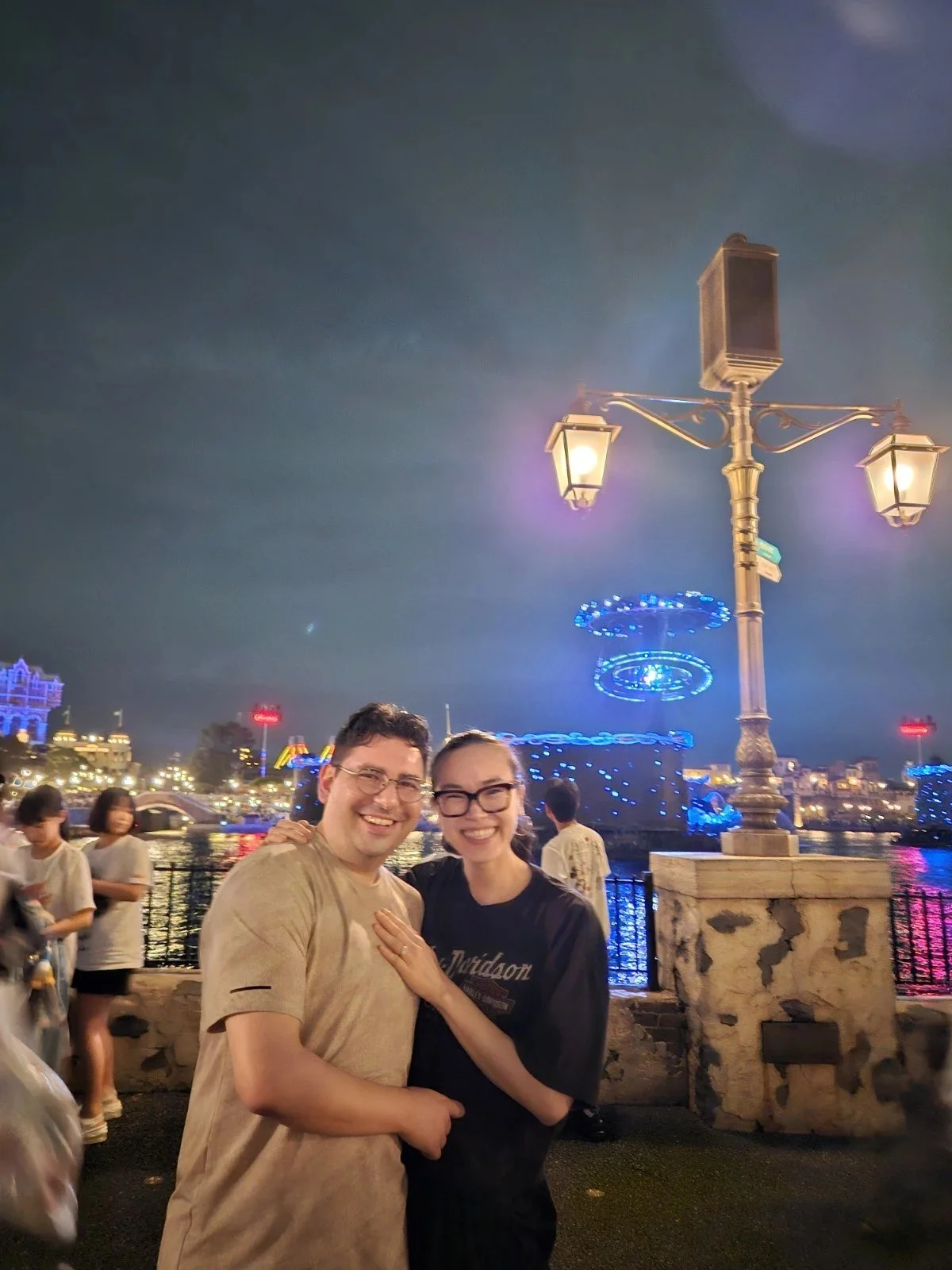 Smiling man and woman standing close together at night near a river, illuminated by decorative street lamps, with colorful lights and a cityscape in the background.