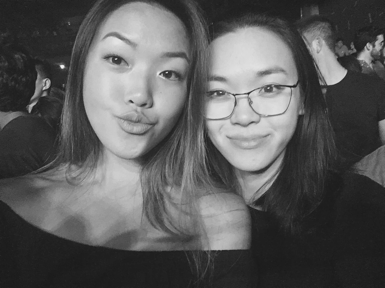 Two young women taking a selfie together at a crowded event or party, one wearing glasses and both smiling.