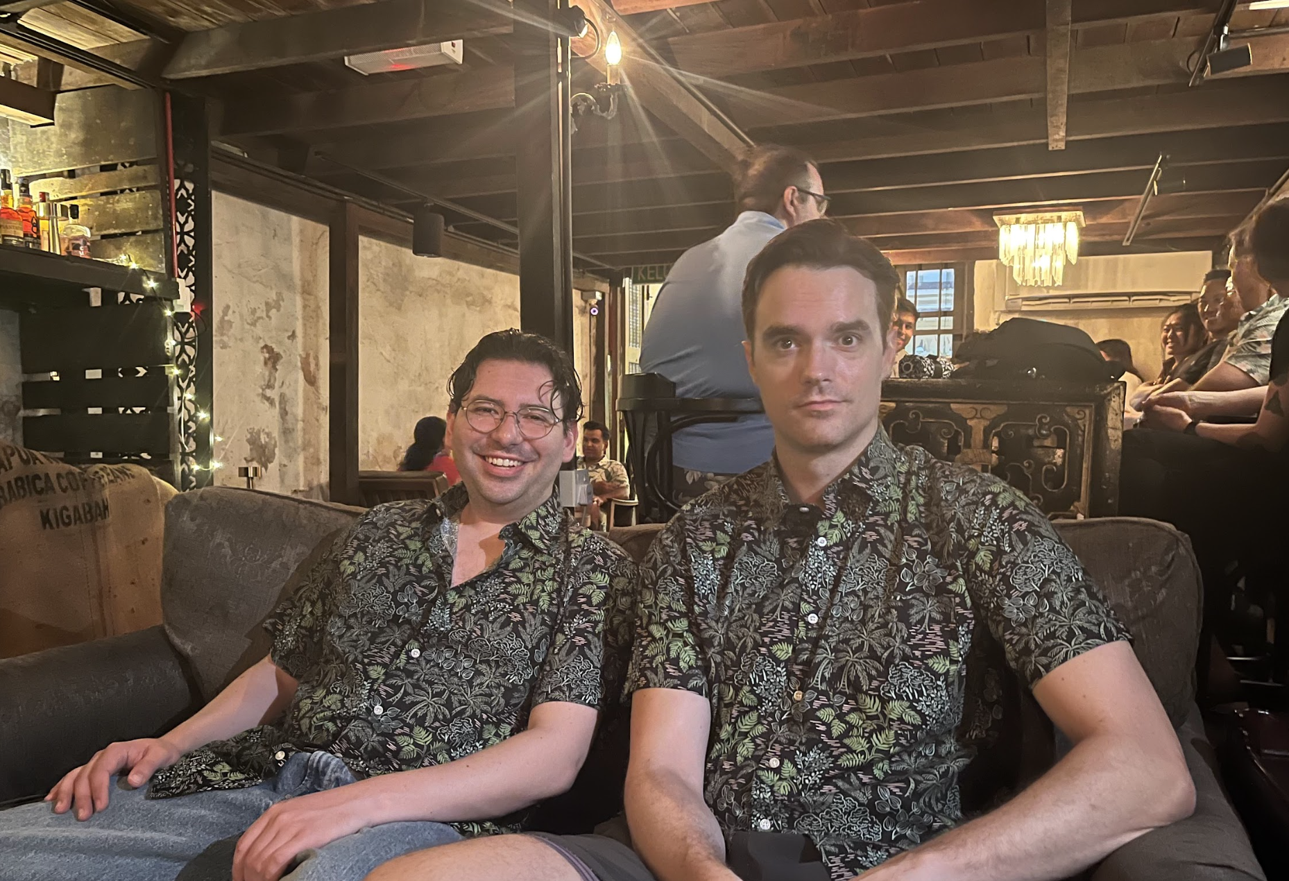 Two men sitting on a sofa in a lively indoor bar or restaurant, both wearing matching black floral shirts. The man on the left is smiling and wearing glasses, while the man on the right has a neutral expression. The background includes other patrons,