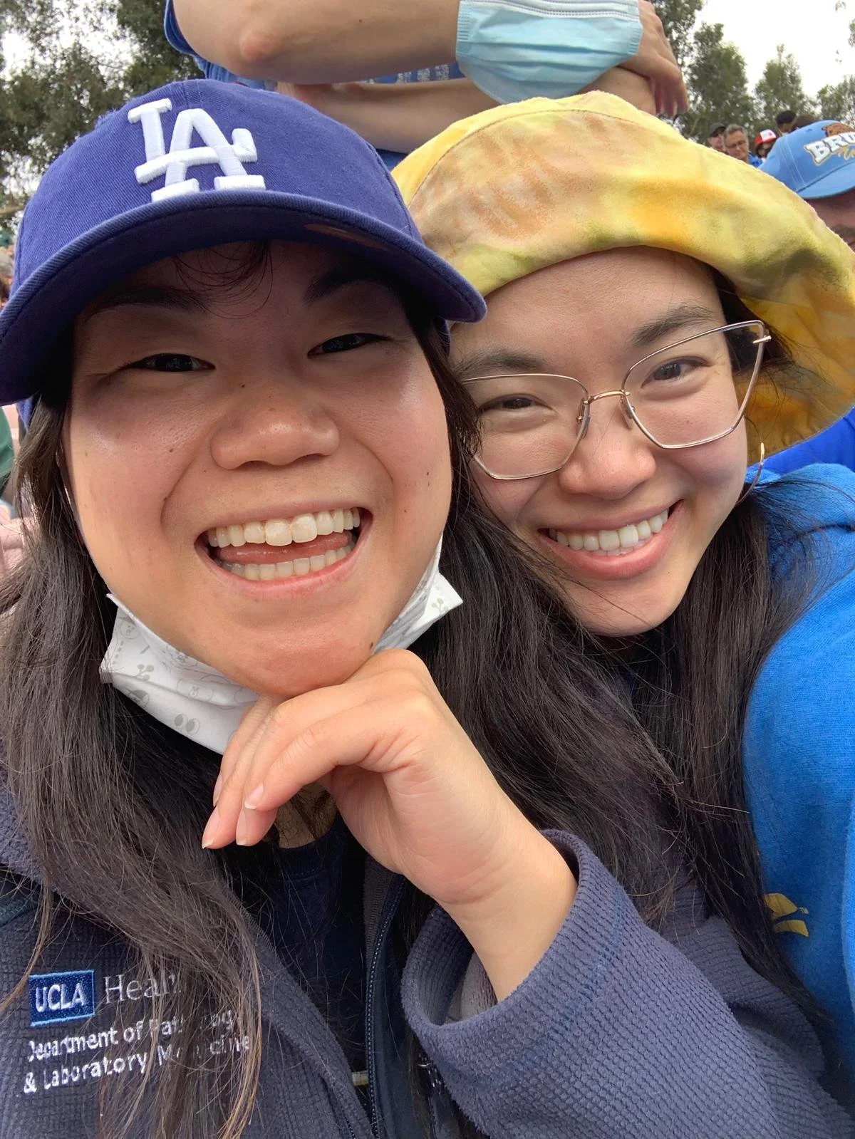 Two women smiling for a selfie outdoors, one wearing a blue UCLA cap and a jacket with UCLA Health logo, and the other wearing glasses and a yellow hat with a colorful pattern.