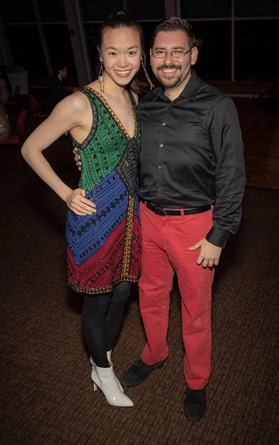 A man and woman pose together smiling at an indoor event, with other people and large windows in the background.