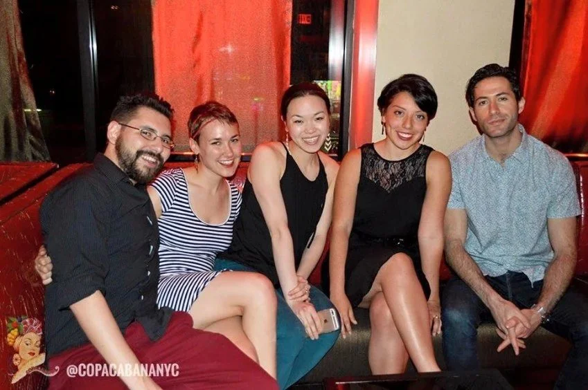 Five friends sitting together on a red booth in a restaurant or bar, smiling at the camera.