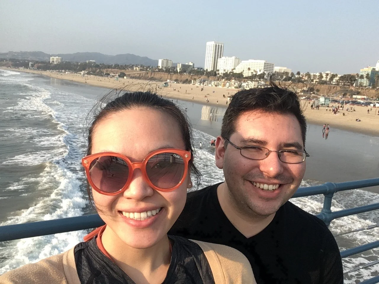 A smiling couple taking a selfie on a boat or pier with a beach and city skyline in the background.