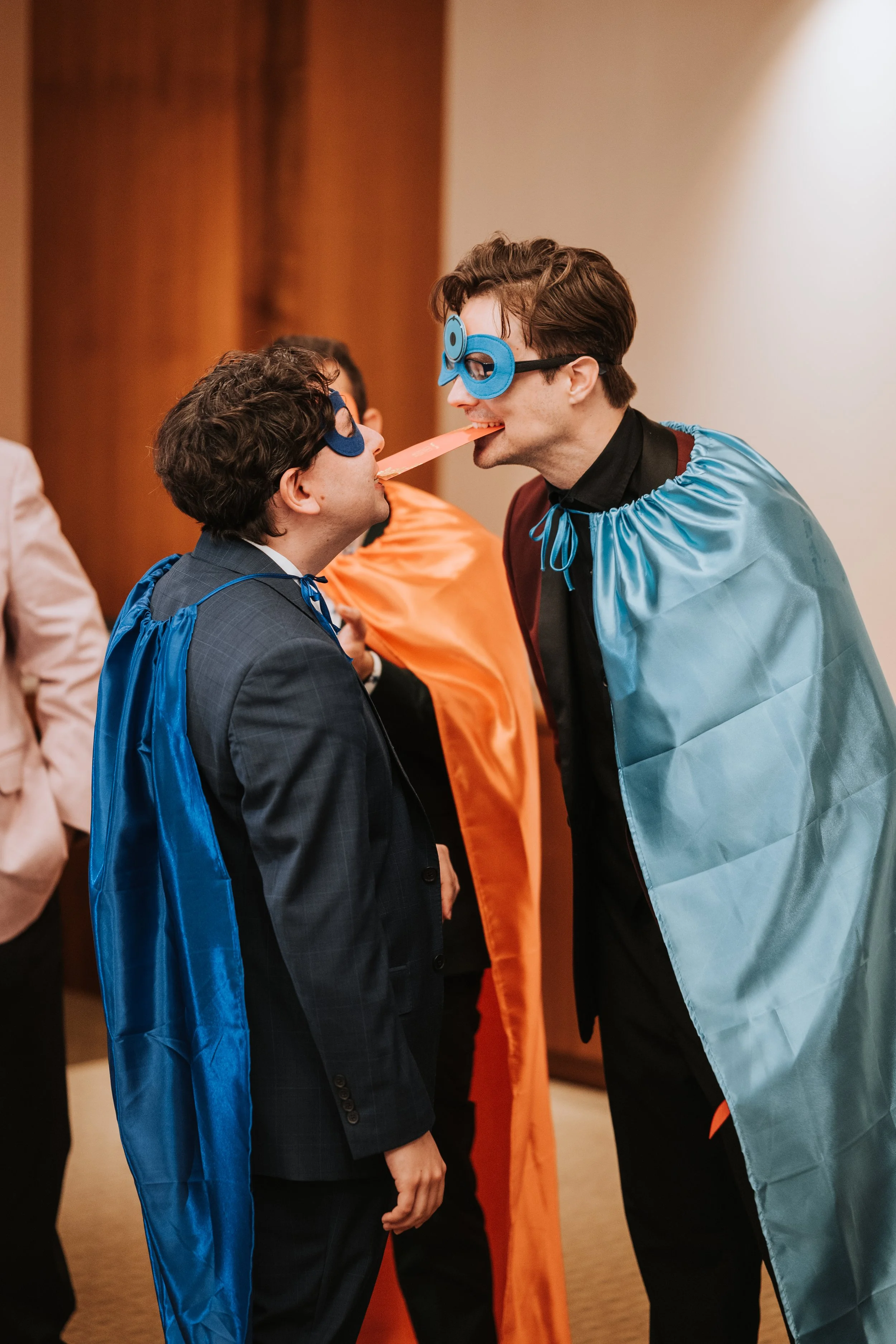 Two men with fun glasses and capes playfully feed each other at an indoor event.