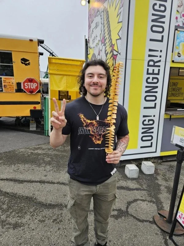 A man with long dark curly hair, wearing a black Iron Maiden t-shirt, stands outdoors in front of a food truck. He is smiling and making a peace sign with his right hand while holding a skewer of spiral-cut fries in his left hand. The background incl