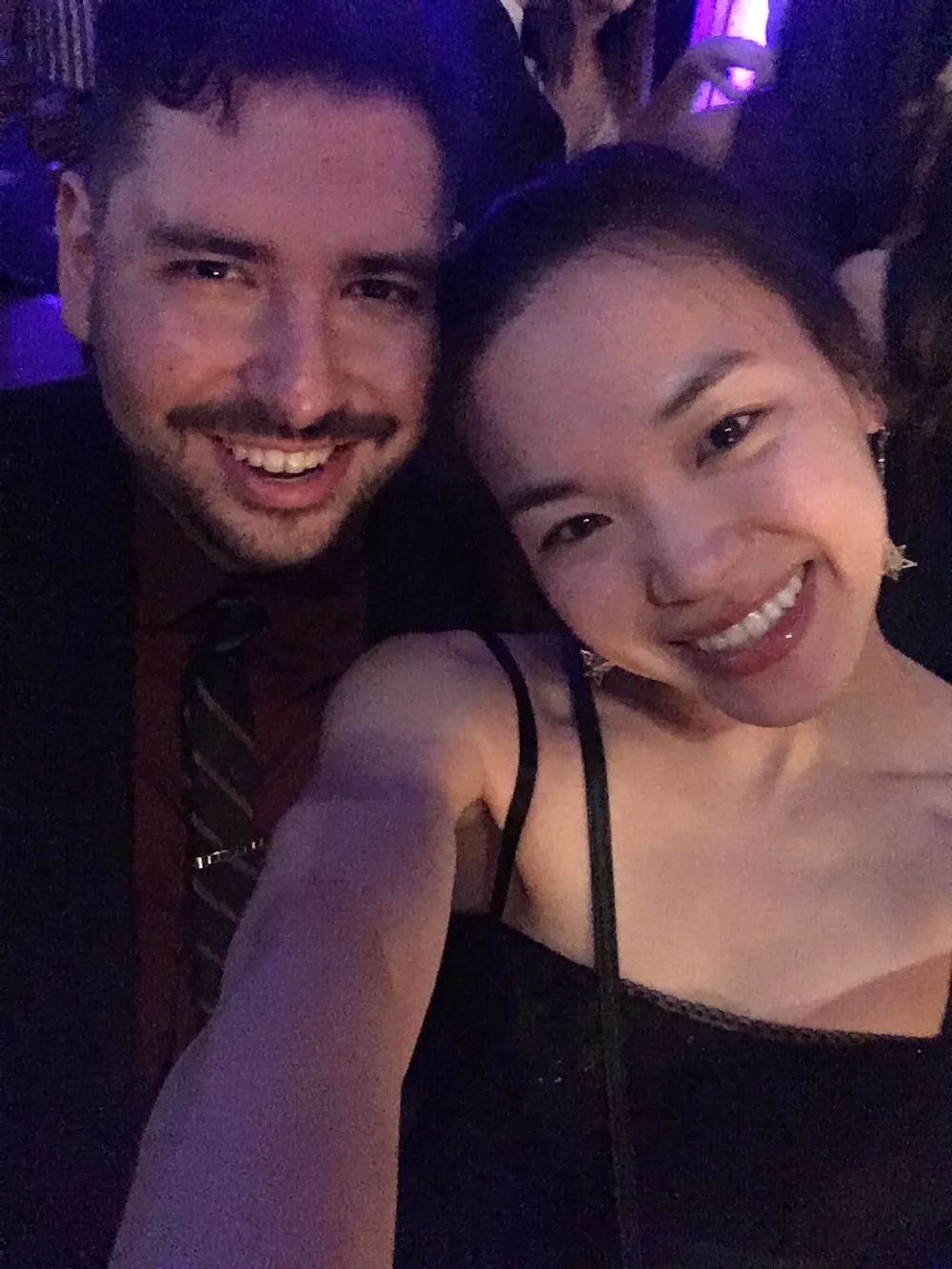 A smiling man and woman taking a selfie together at a party or event with low lighting and purple hues.