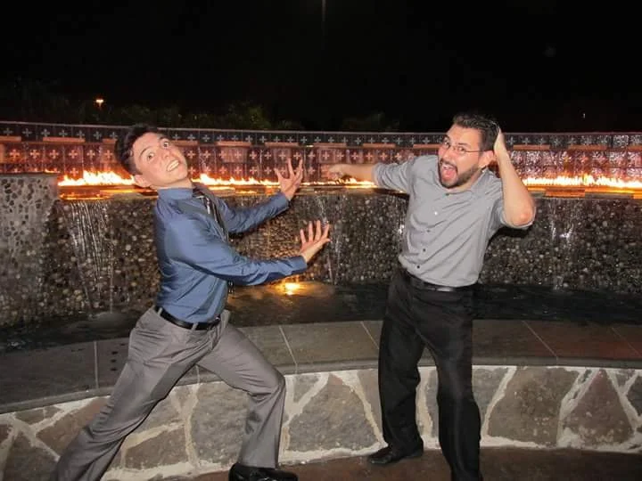 Two men in dress shirts and dress pants playfully posing in front of a fountain with fire behind them at night.
