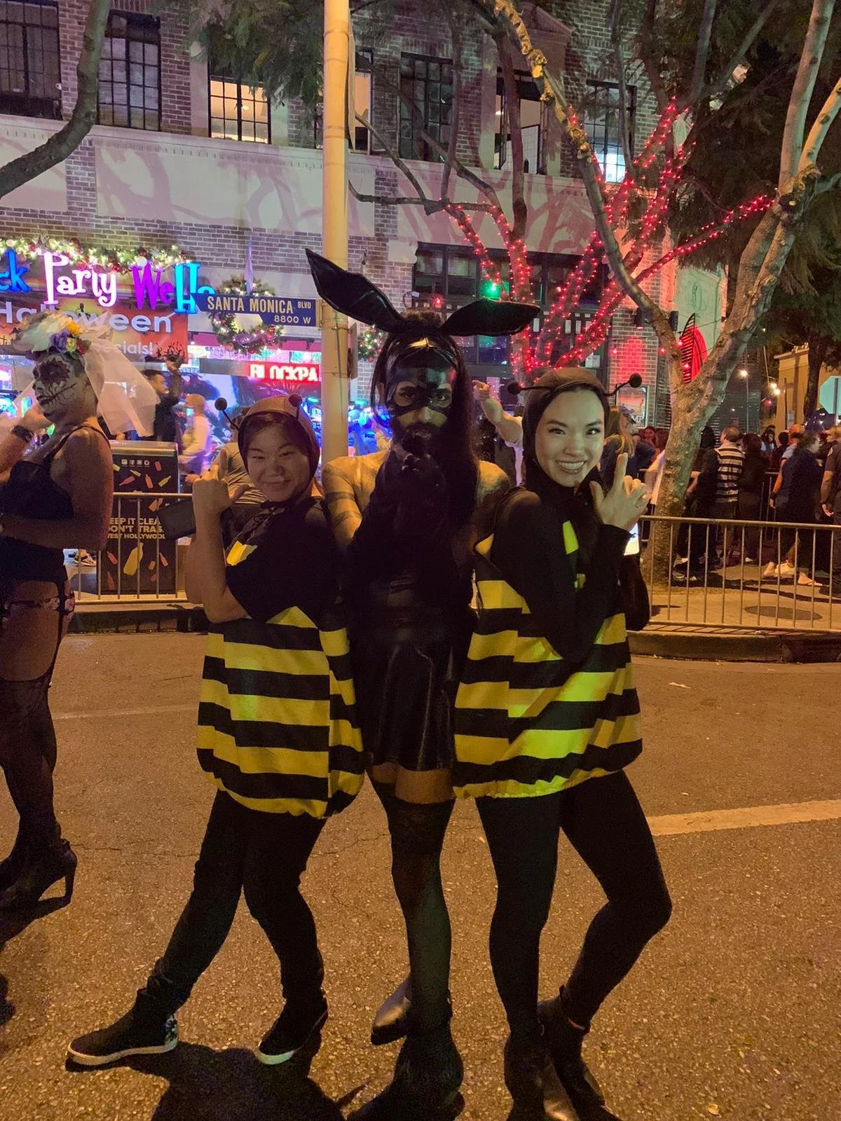 Three women in Halloween costumes posing on a street at night during a Halloween celebration. Two women are dressed as bees, and the third woman is dressed as a black cat with bunny ears and black face paint.