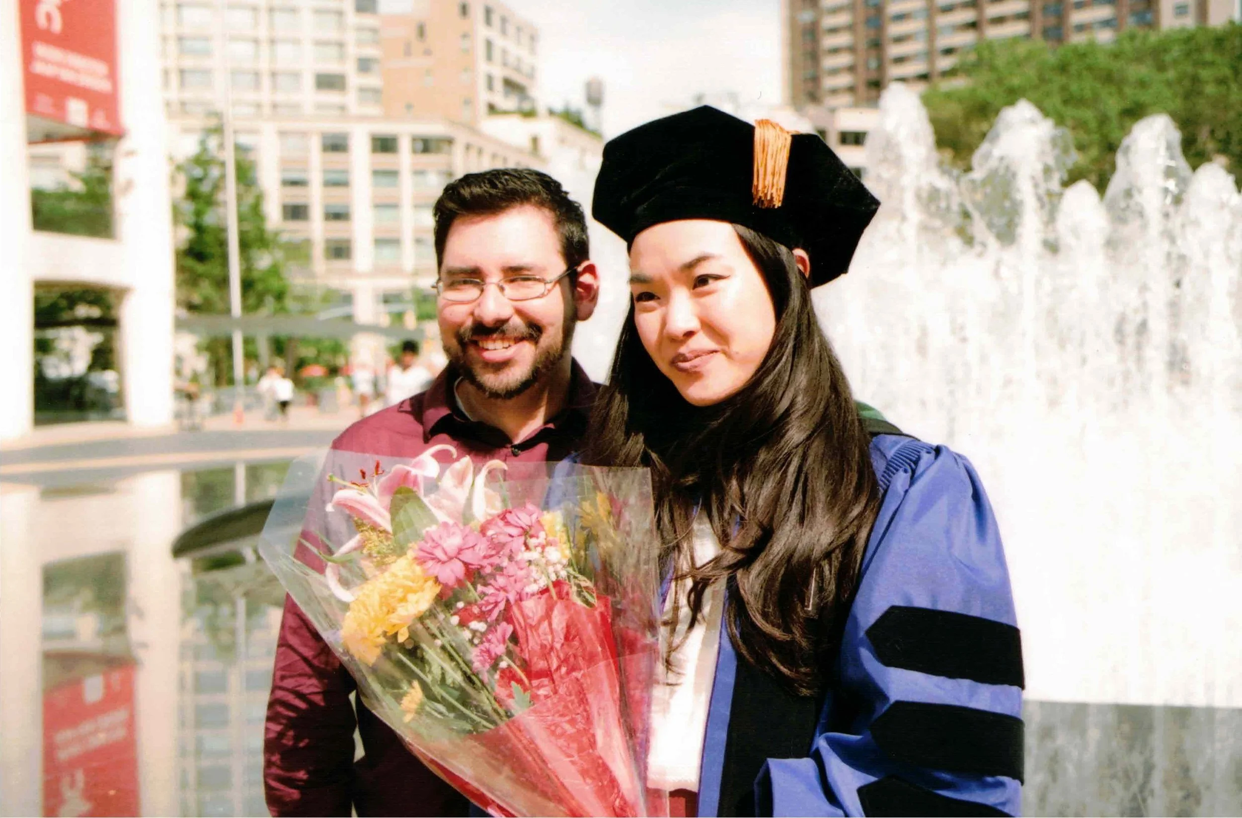 A young woman in graduation attire holding a bouquet of flowers, standing next to a man on an urban campus with a fountain and buildings in the background.