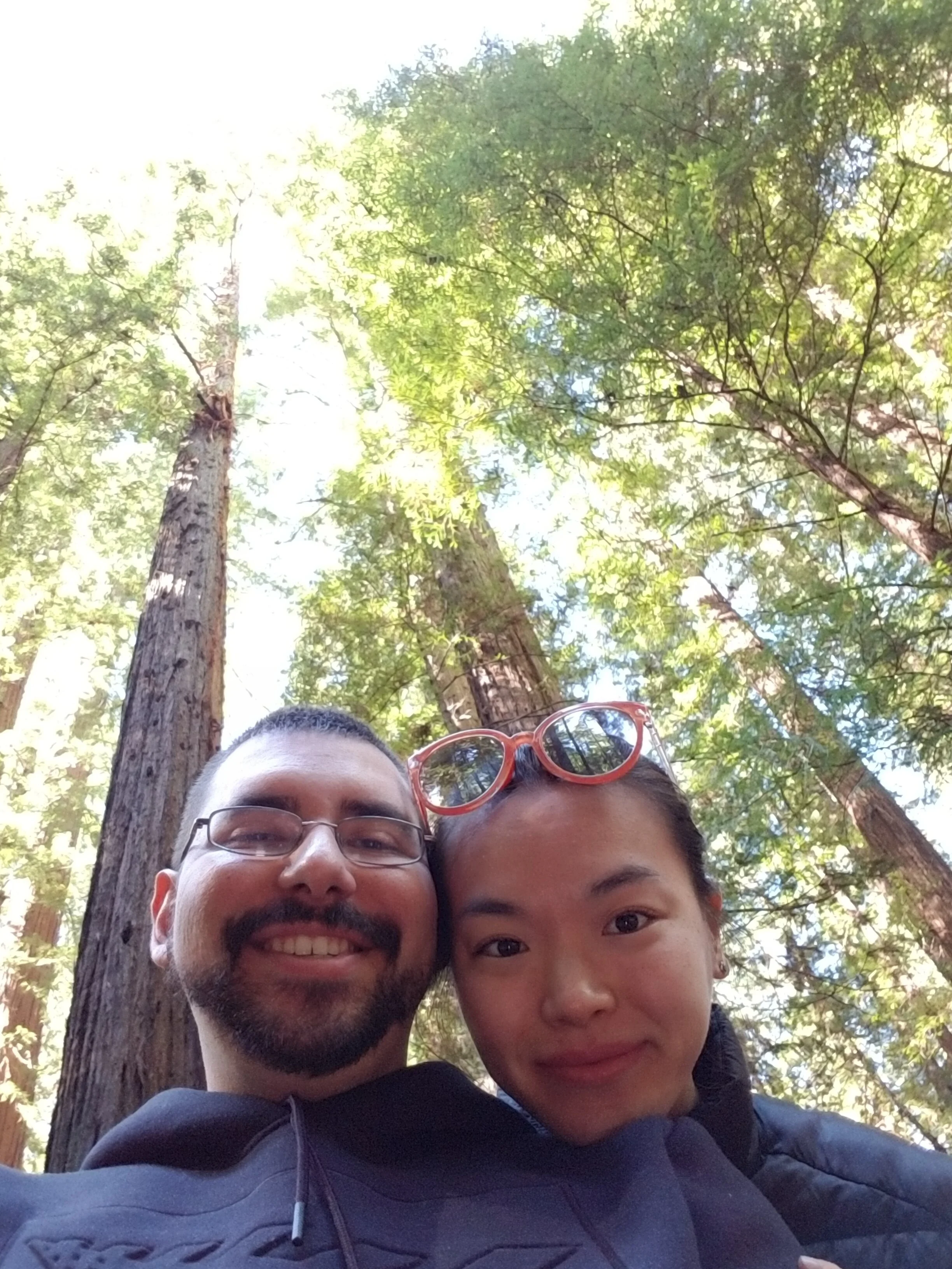 Smiling man and woman taking a selfie together outdoors in a forest with tall trees and sunlight filtering through the green leaves.
