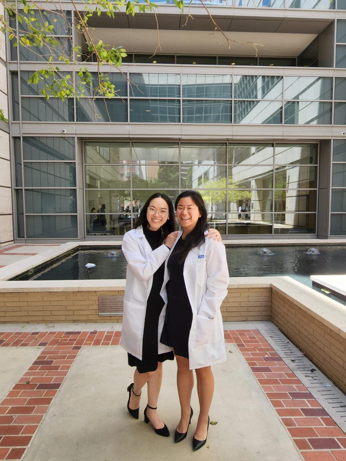 Two women in black dresses and white lab coats standing outside in front of a modern glass building with a fountain, smiling and laughing.