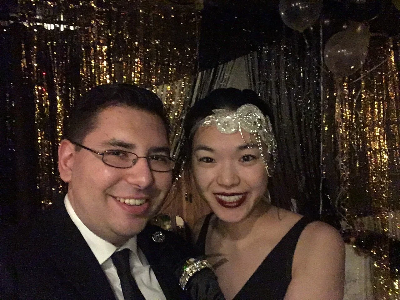 A man and woman smiling, dressed in formal attire at a party with a gold sequin backdrop. The woman is wearing a black dress and a silver headpiece. The man is wearing glasses, a black suit, and a white shirt.