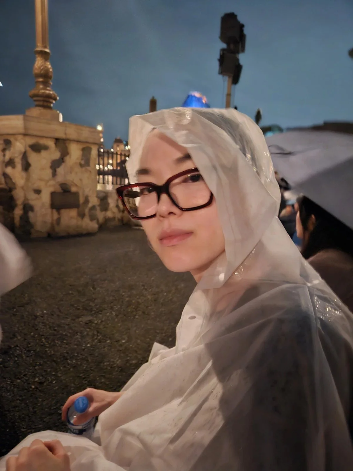 A person wearing glasses and a clear plastic raincoat sitting outdoors at night near a stone fence with a blue light in the background.