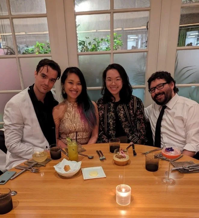 Four people sitting at a restaurant table with drinks and desserts, smiling for the photo, with a large window behind them showing plants outside.