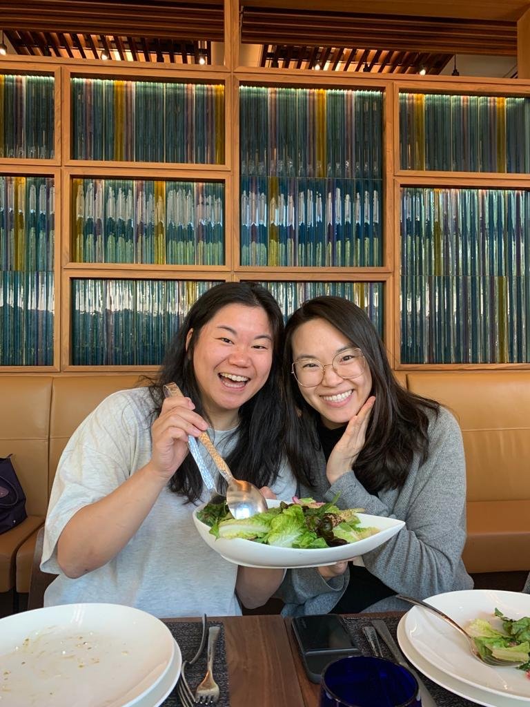 Two women smiling and holding a large salad at a restaurant table with empty plates, utensils, a phone, and a glass, in front of a bookshelf filled with colorful books.