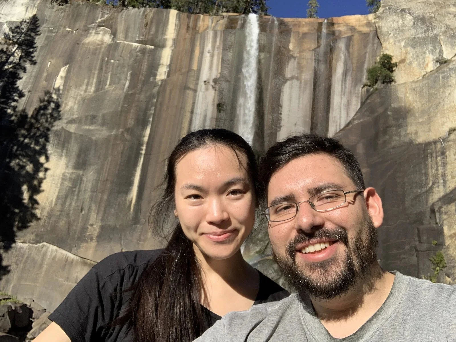 A smiling couple taking a selfie in front of a waterfall at a national park.
