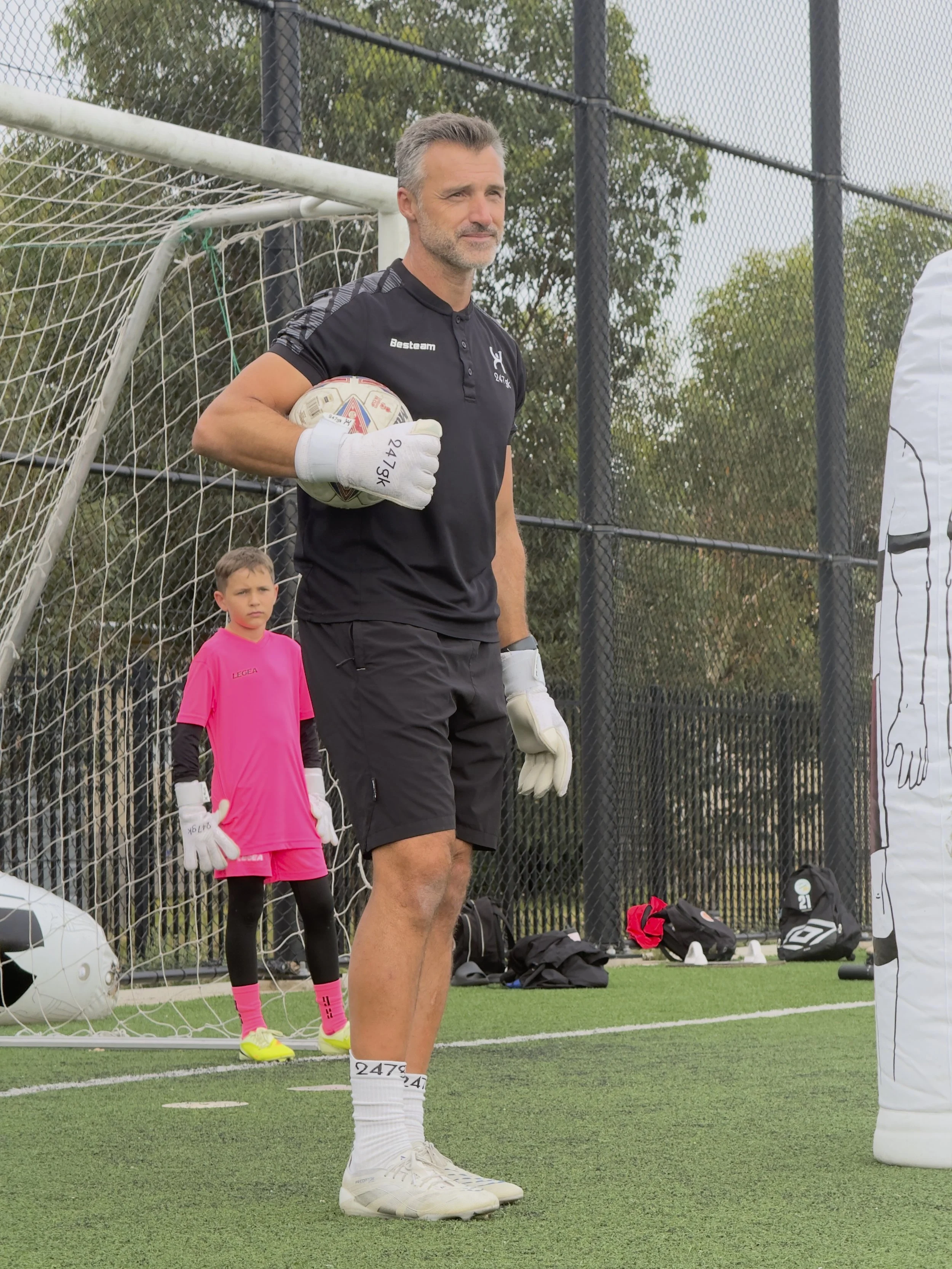 A middle-aged man in black clothing and white soccer gloves holds a soccer ball next to a goalpost on a soccer field. A young goalie in pink uniform stands behind him, also wearing gloves, on the field.