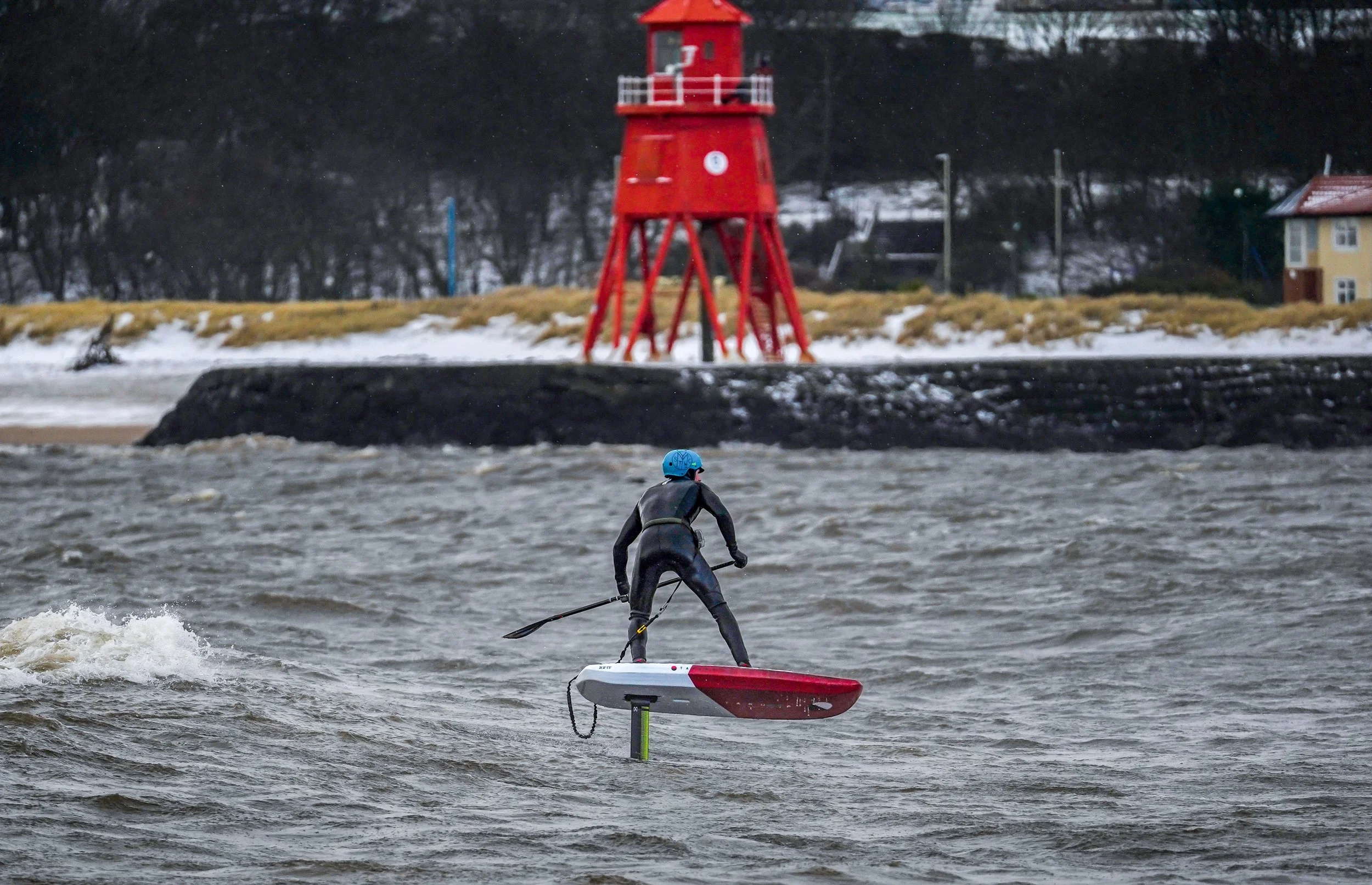 Person in wetsuit and blue helmet riding a hydrofoil board on choppy water with a red lighthouse and snow-covered shoreline in the background.
