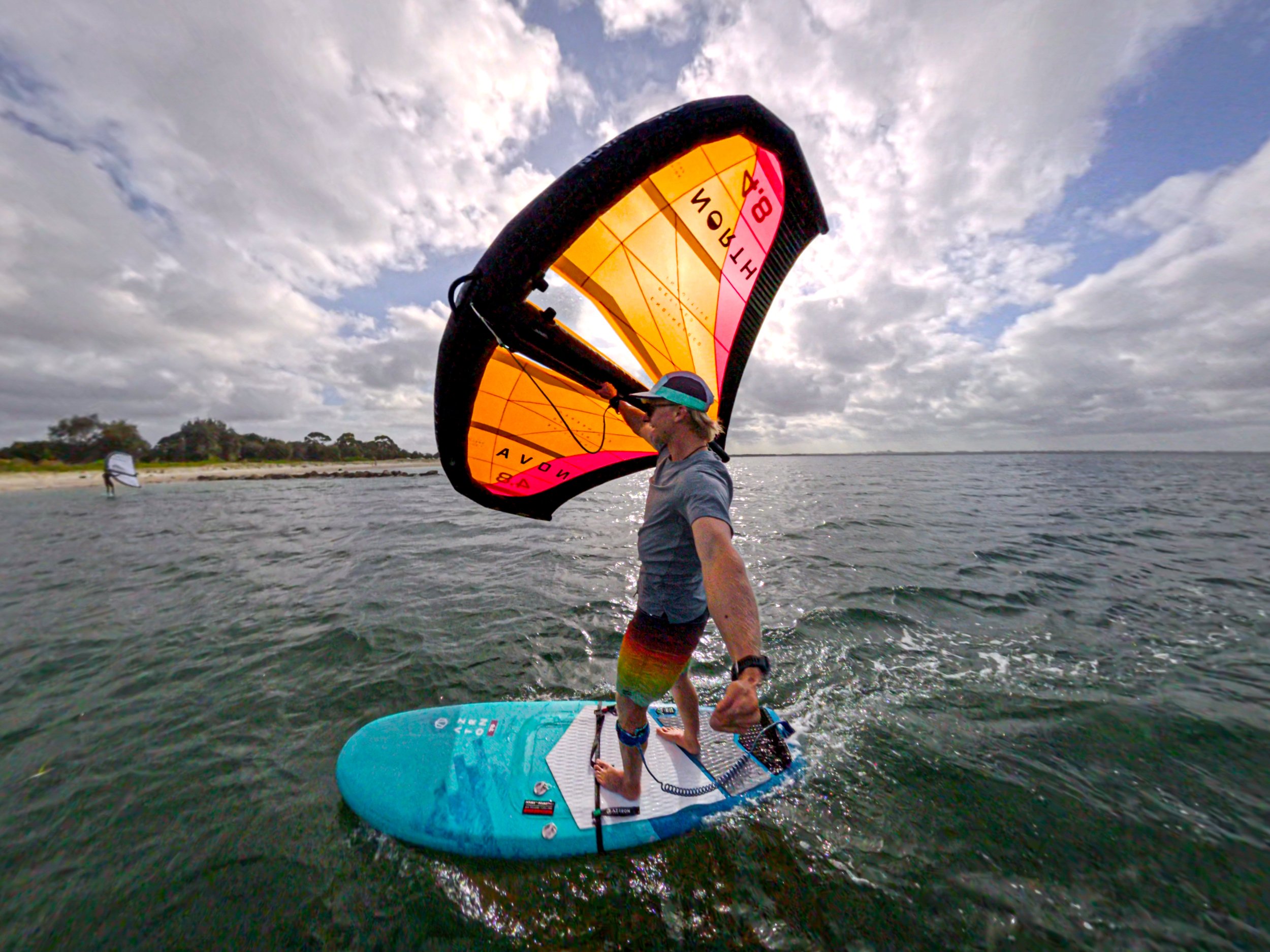 Man wingsurfing on a blue board with go-pro camera, holding onto an orange and pink sail, with a second windsurfer in the background near a beach under a partly cloudy sky.
