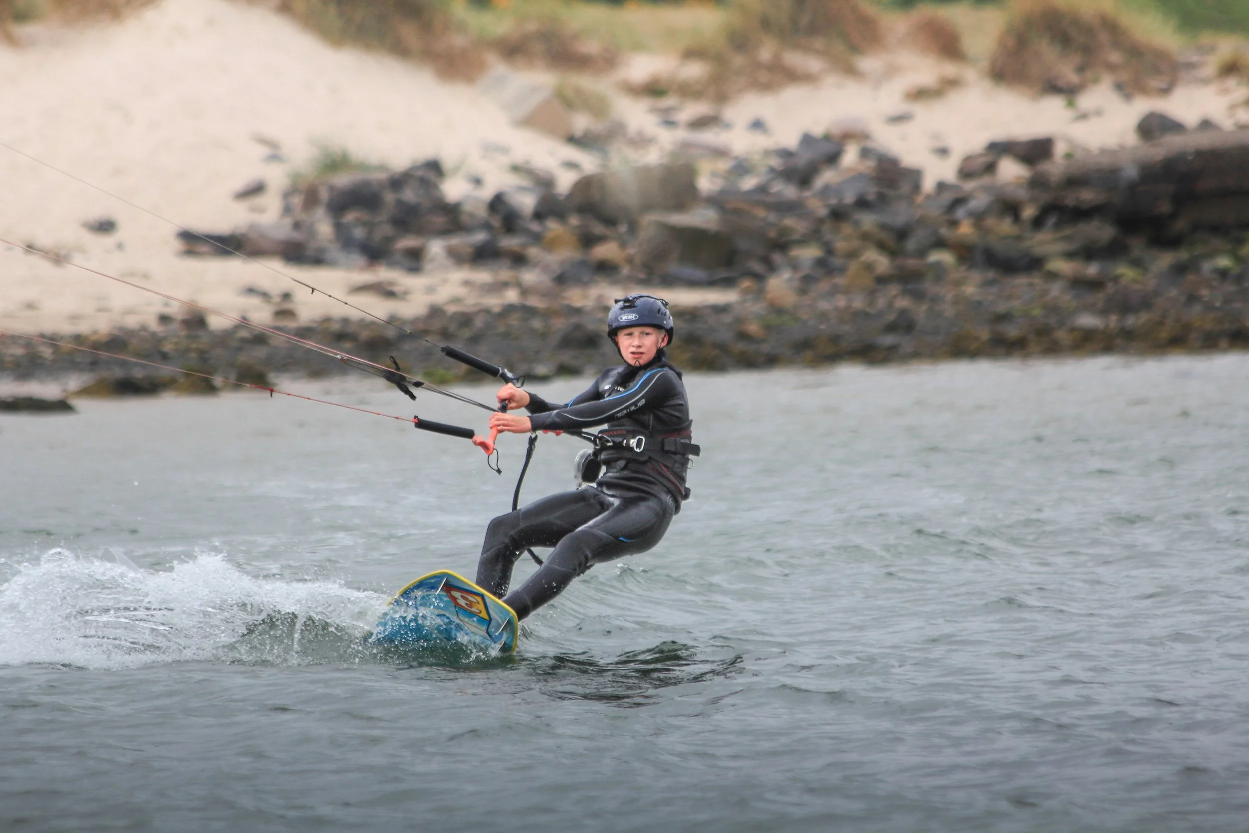 A person wearing a helmet and wetsuit is kiteboarding on a body of water near a rocky shoreline.