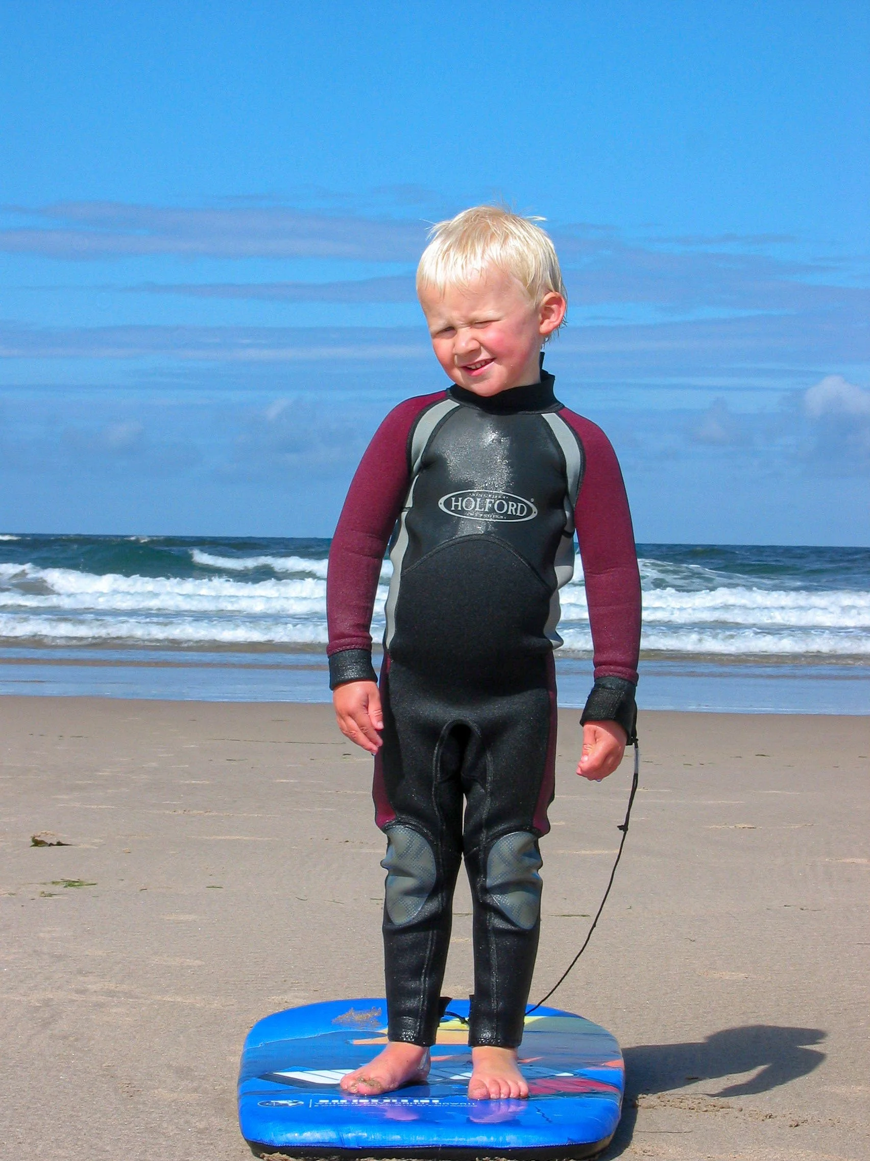 A young boy in a wetsuit standing on a surfboard on a sandy beach with ocean waves and a partly cloudy sky in the background.