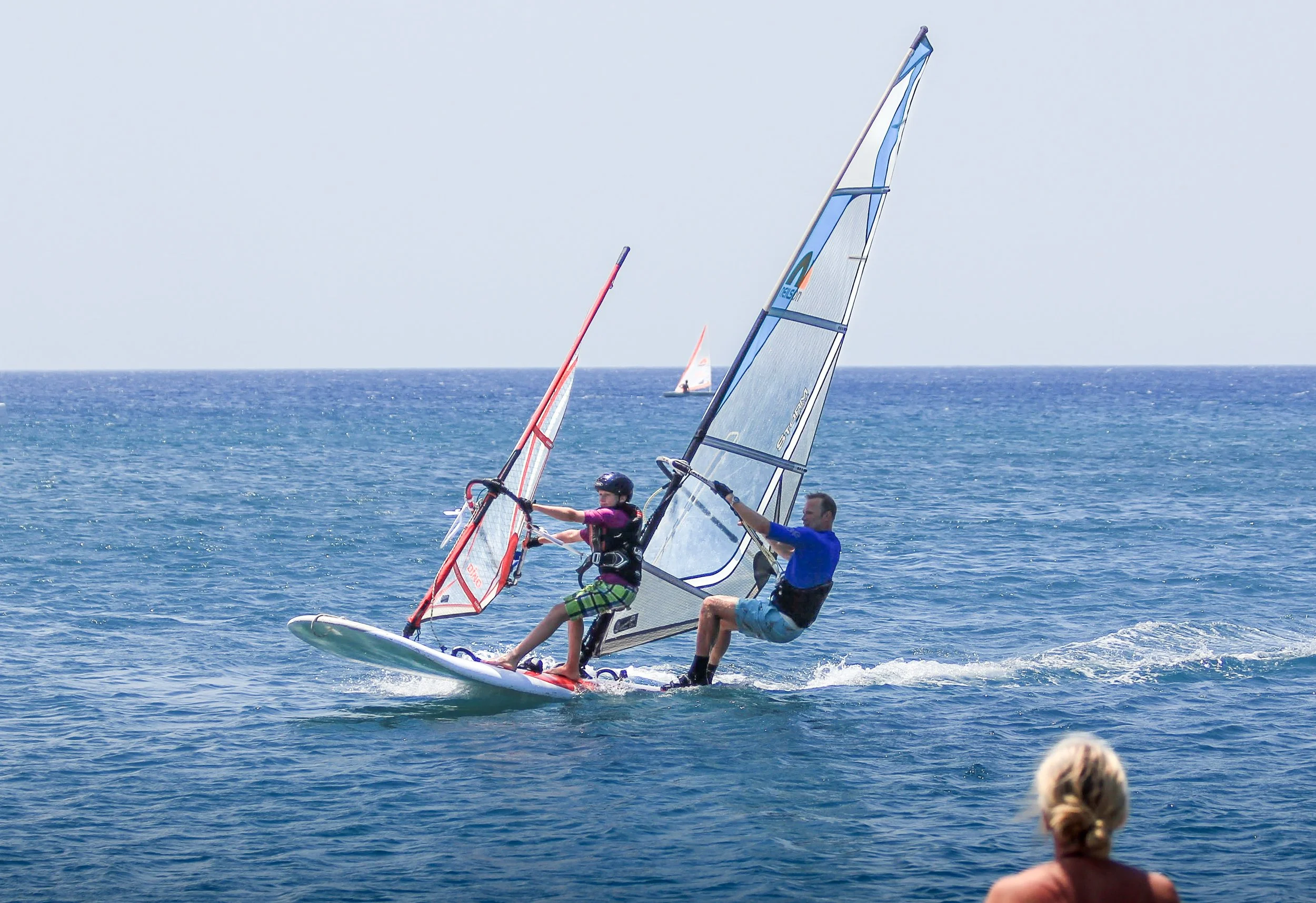 Two people windsurfing on the ocean with a small sailboat in the background and a woman watching from the shore.