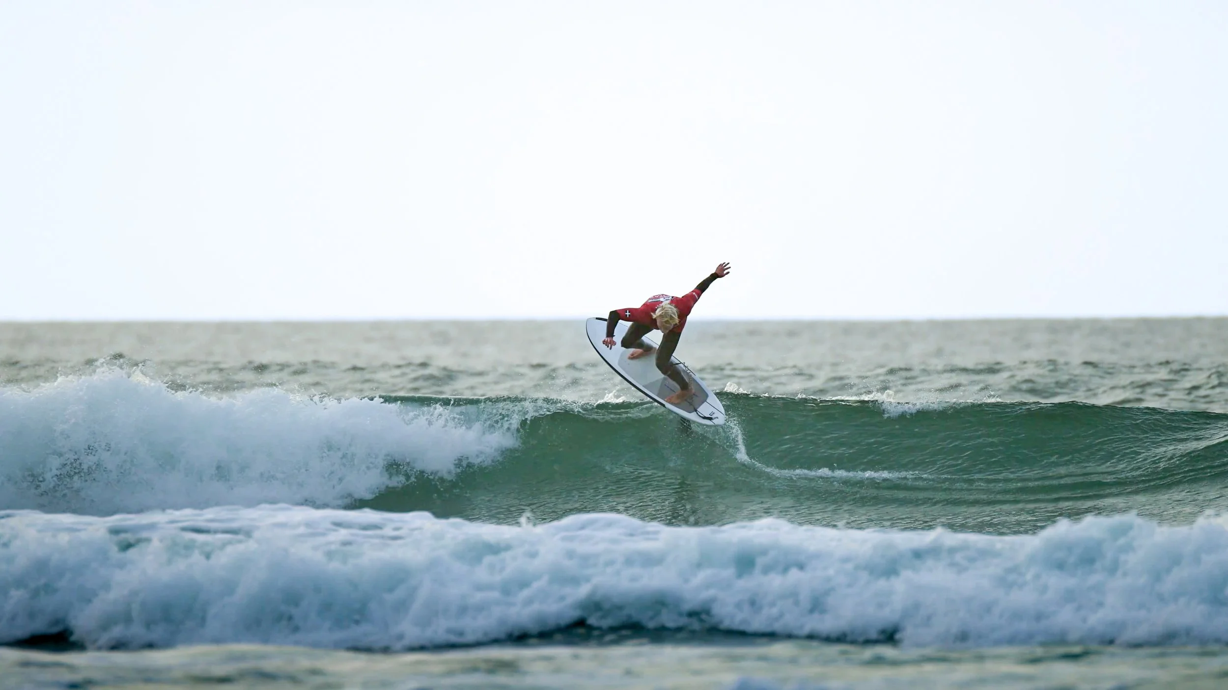 A surfer in red and black competition jersey riding a wave on a hydrofoil.