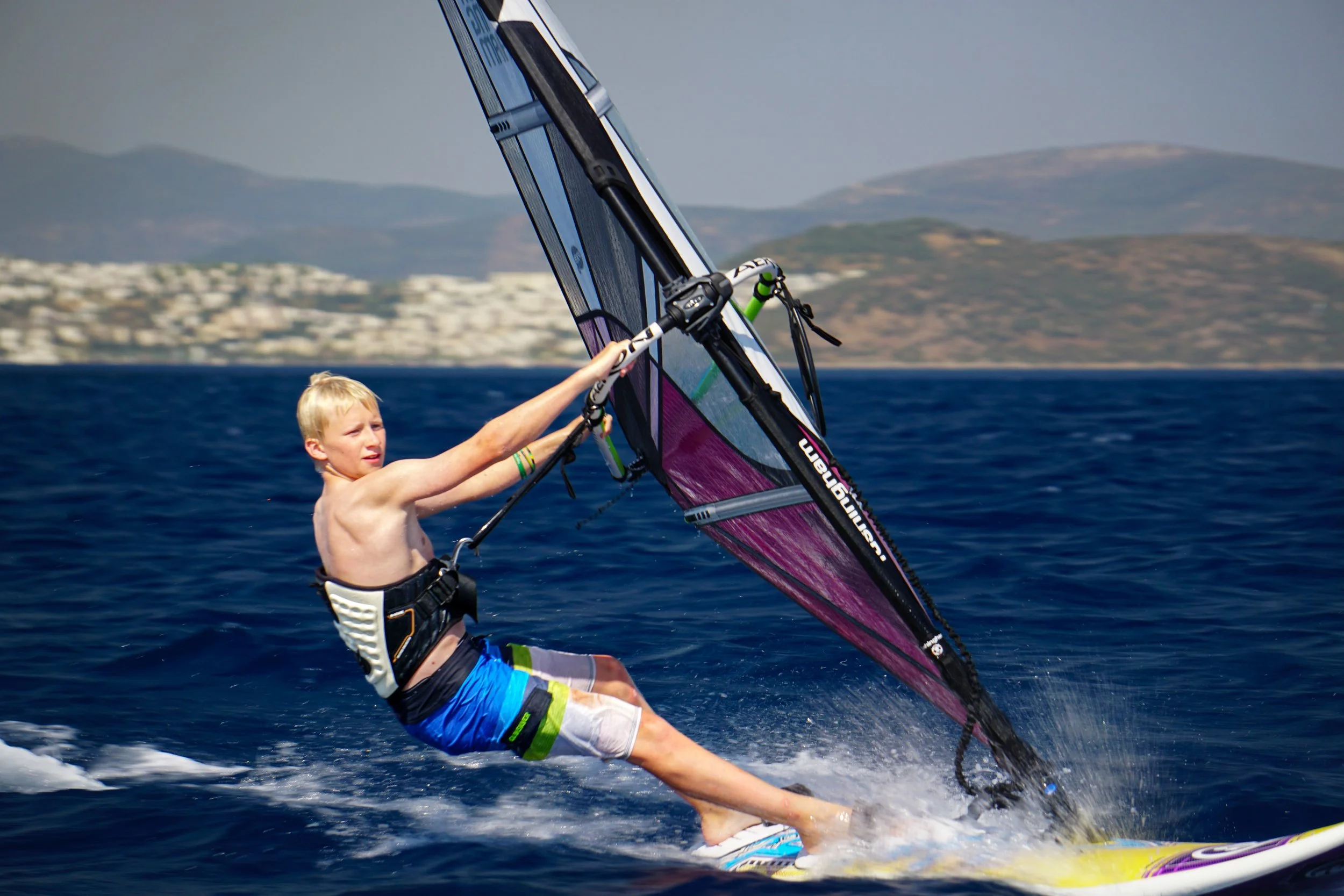 A young boy windsurfing on the ocean with mountains and a distant shoreline in the background.