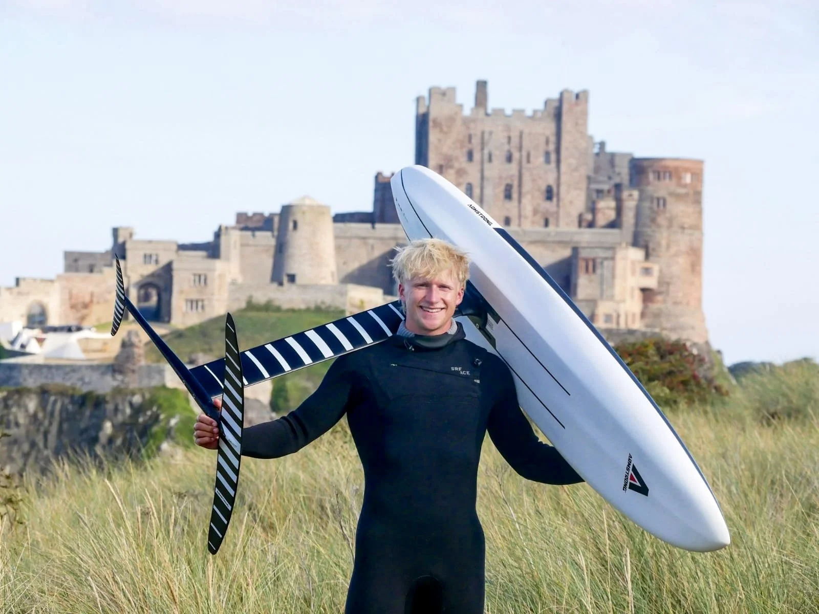 A smiling man in a black wetsuit holding a hydrofoil, standing in a grassy field with a historic castle in the background.