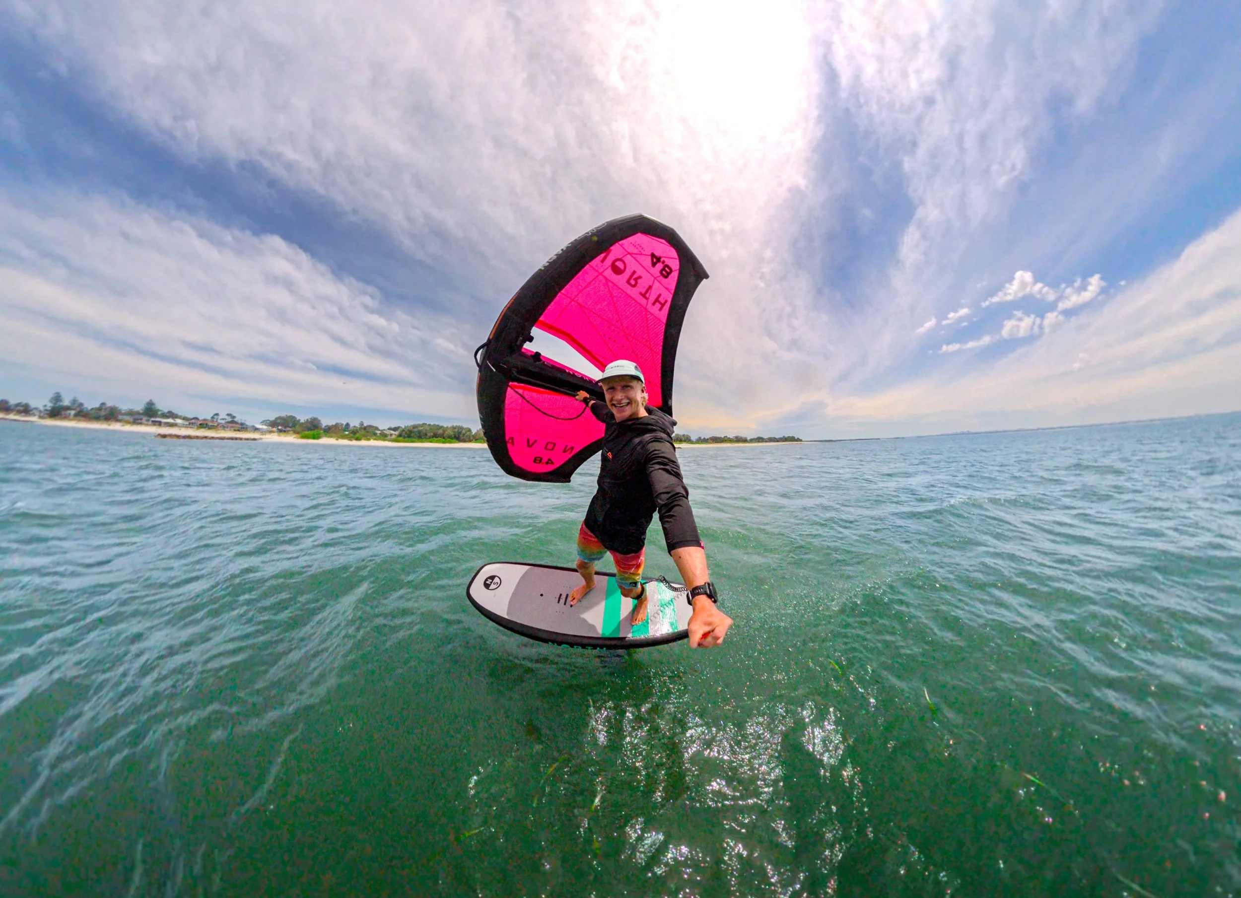A man wingfoiling on the water with a pink wing, smiling, wearing a black jacket, colorful shorts, a white cap, and a watch, with a coastal shoreline and cloudy sky in the background.