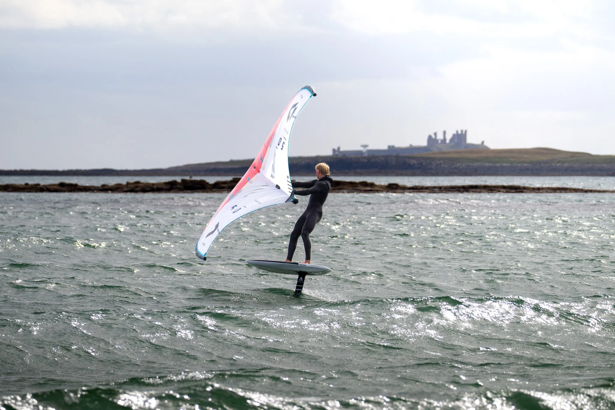Person wearing a wetsuit on a hydrofoil surfing on choppy water with a castle in the background.